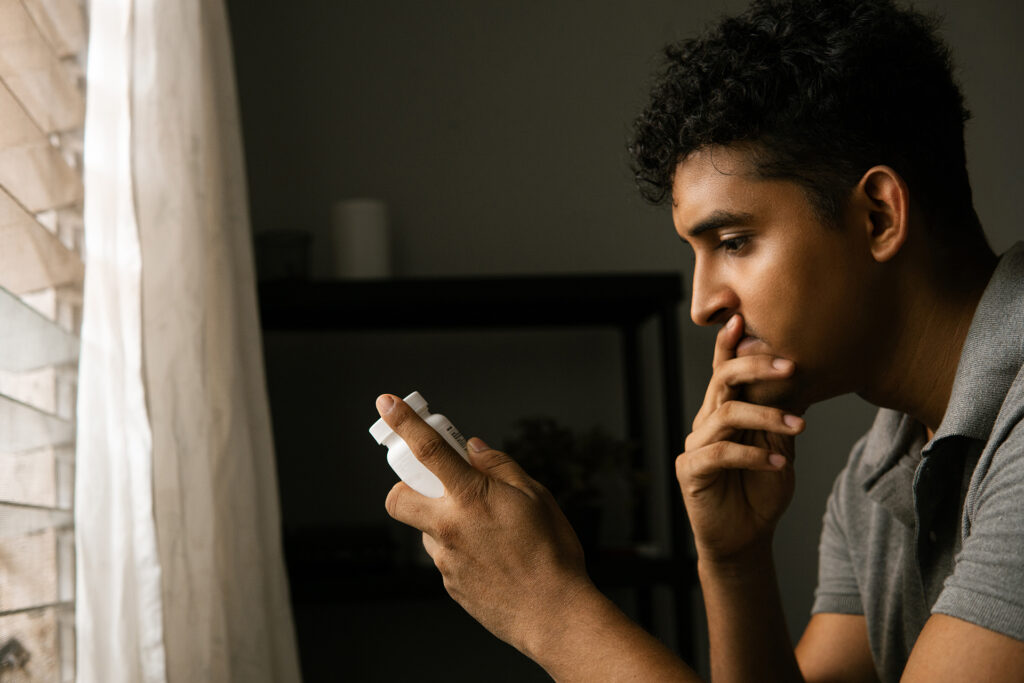 Young man looks at prescription pill bottle in hand in front of a window in a dark bedroom