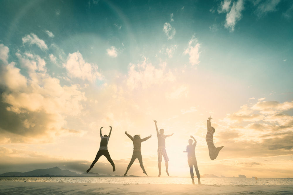Group of five young people leaping triumphantly into the air on a sunlit beach