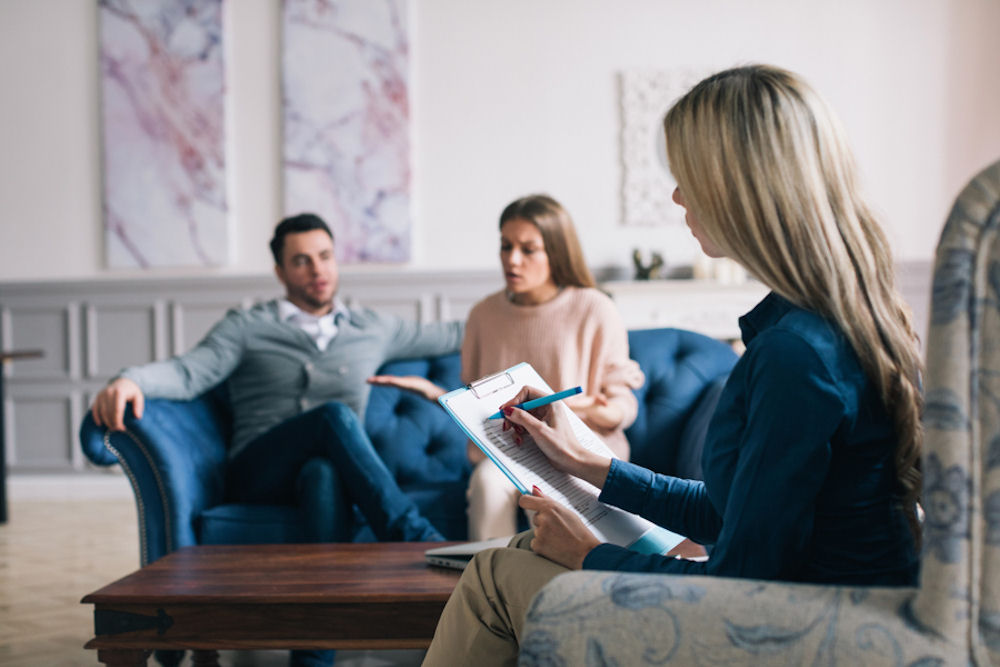 Young therapist taking notes of discussion between young man and young woman on distance couch