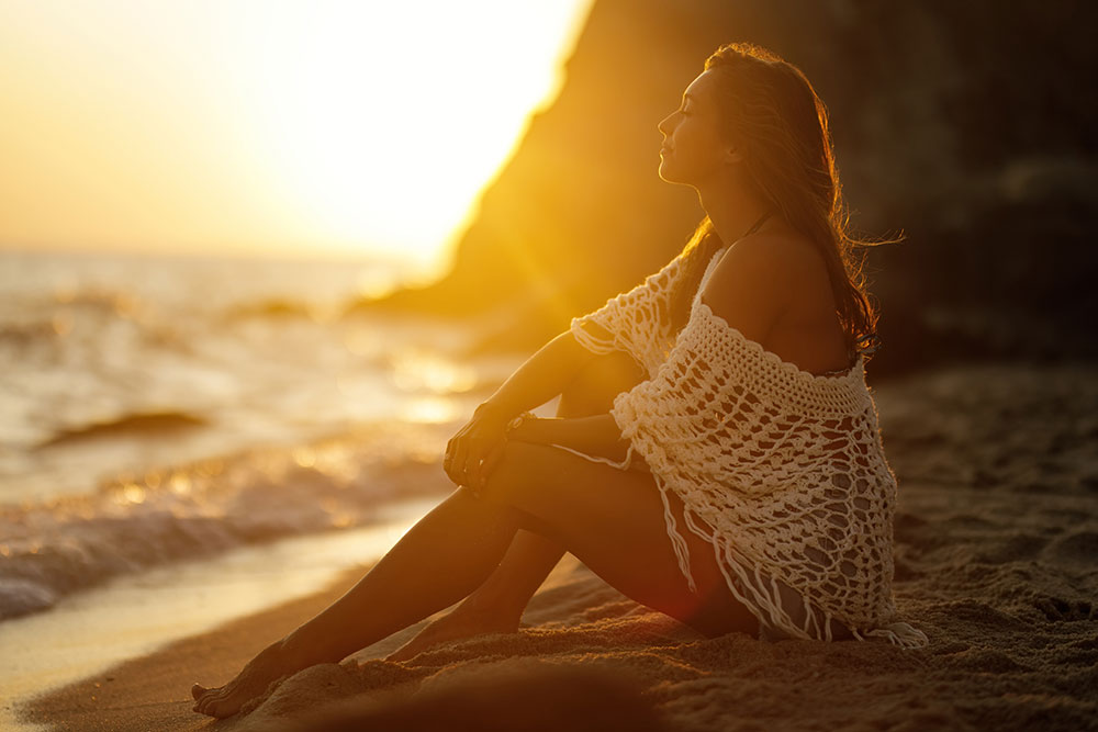 Woman in swimsuit and white woven cover-up, sitting on the beach while sun sets behind Haystack Rock