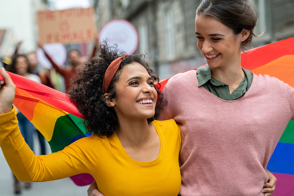 Two women smiling and holding rainbow flag while marching in Pride parade
