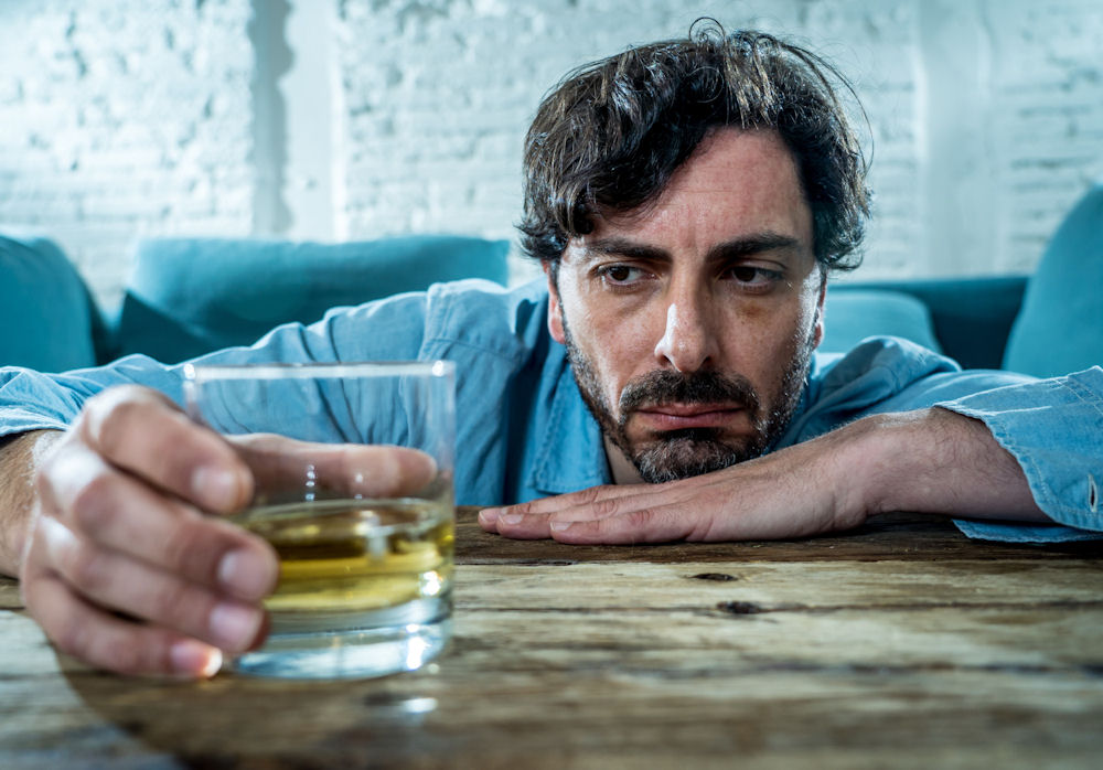 Man stars at glass of whiskey neat while slumped on his hand at an in-home bar