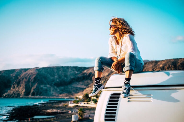 Woman in boho clothing and Converse shoes sitting on top of a VW bus and looking out at the distant ocean