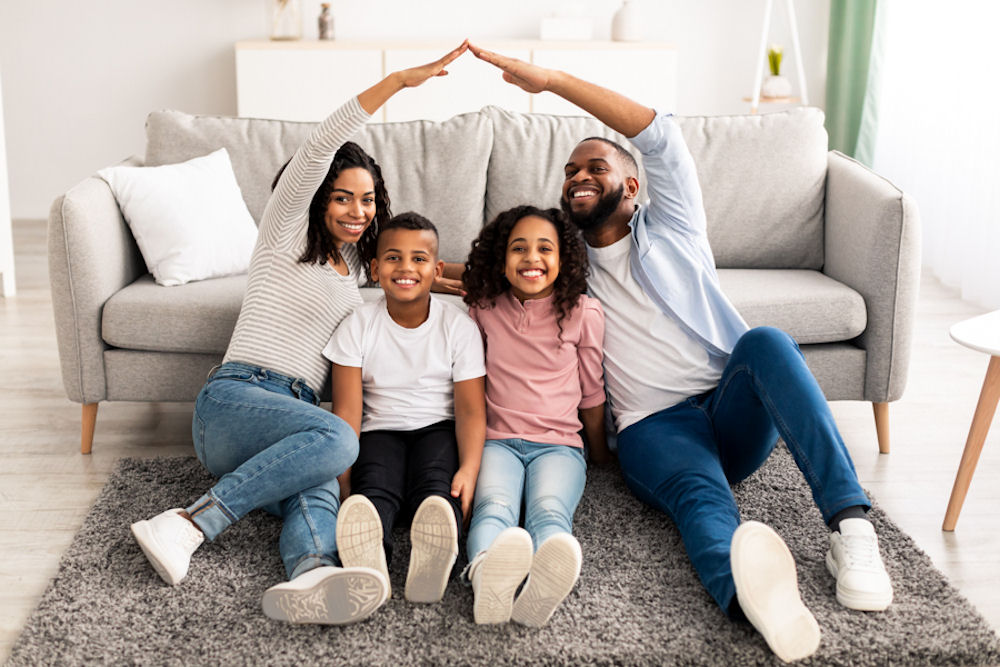 Heterosexual couple reaching up to form arch above son and daughter, while all sit on grey rug in front of grey couch