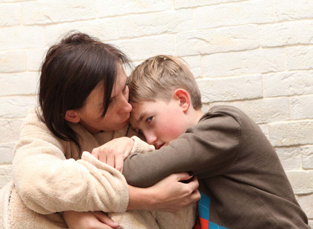 Alcoholic mother wearing fleece robe kissing son who sneaks look at camera