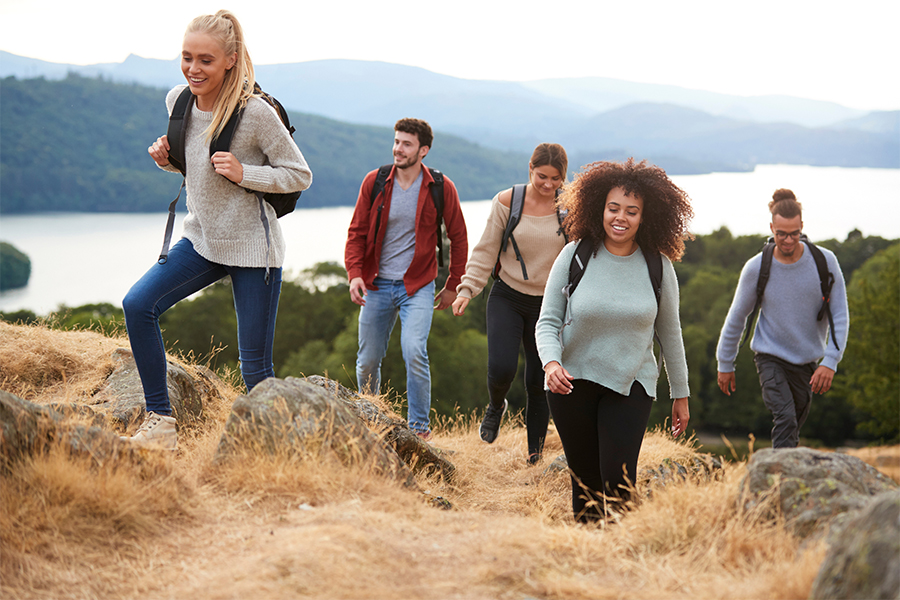 Group of friends of different genders hiking over hill with lake, mountains, and forest in the background