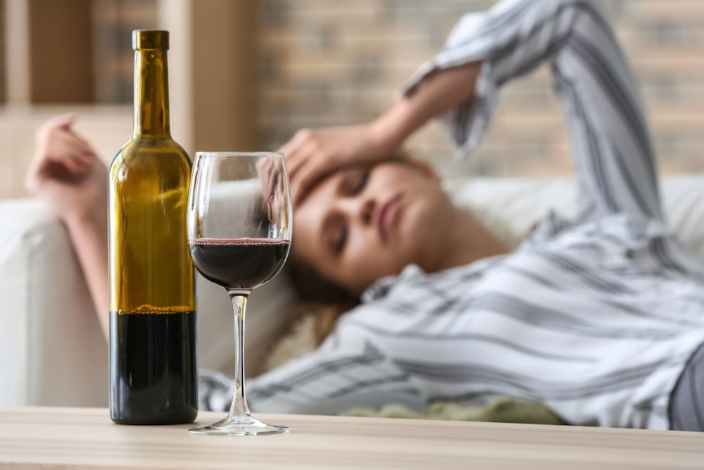 Woman lying on couch holding head next to stemmed glass and wine bottle, each one-third full of red wine