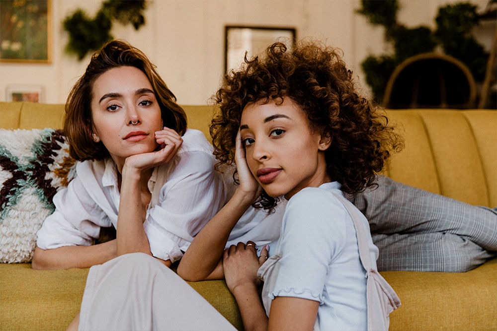 Two young woman lounging on and near a golden rod couch in tousled clothing