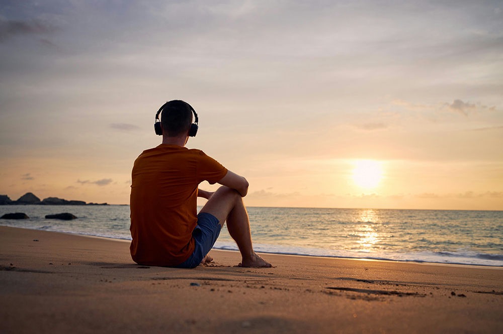 Man sitting on beach wearing can-style headphones watching the sun set