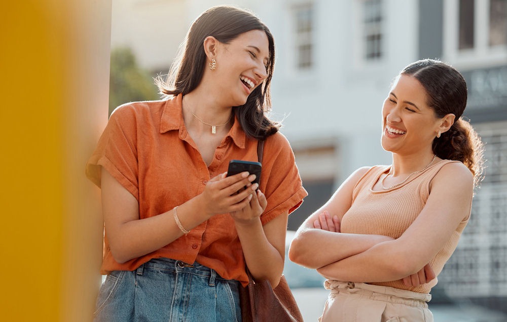 Young women laughing together in outdoor hallway while looking at smartphone